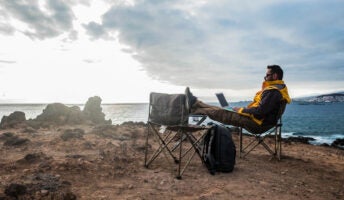 Man in rugged jacket works remotely on laptop sitting on cliff