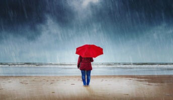 Person standing with an umbrella in the rain on a beach.