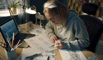 An artist drawing at her desk with a computer in front of her.